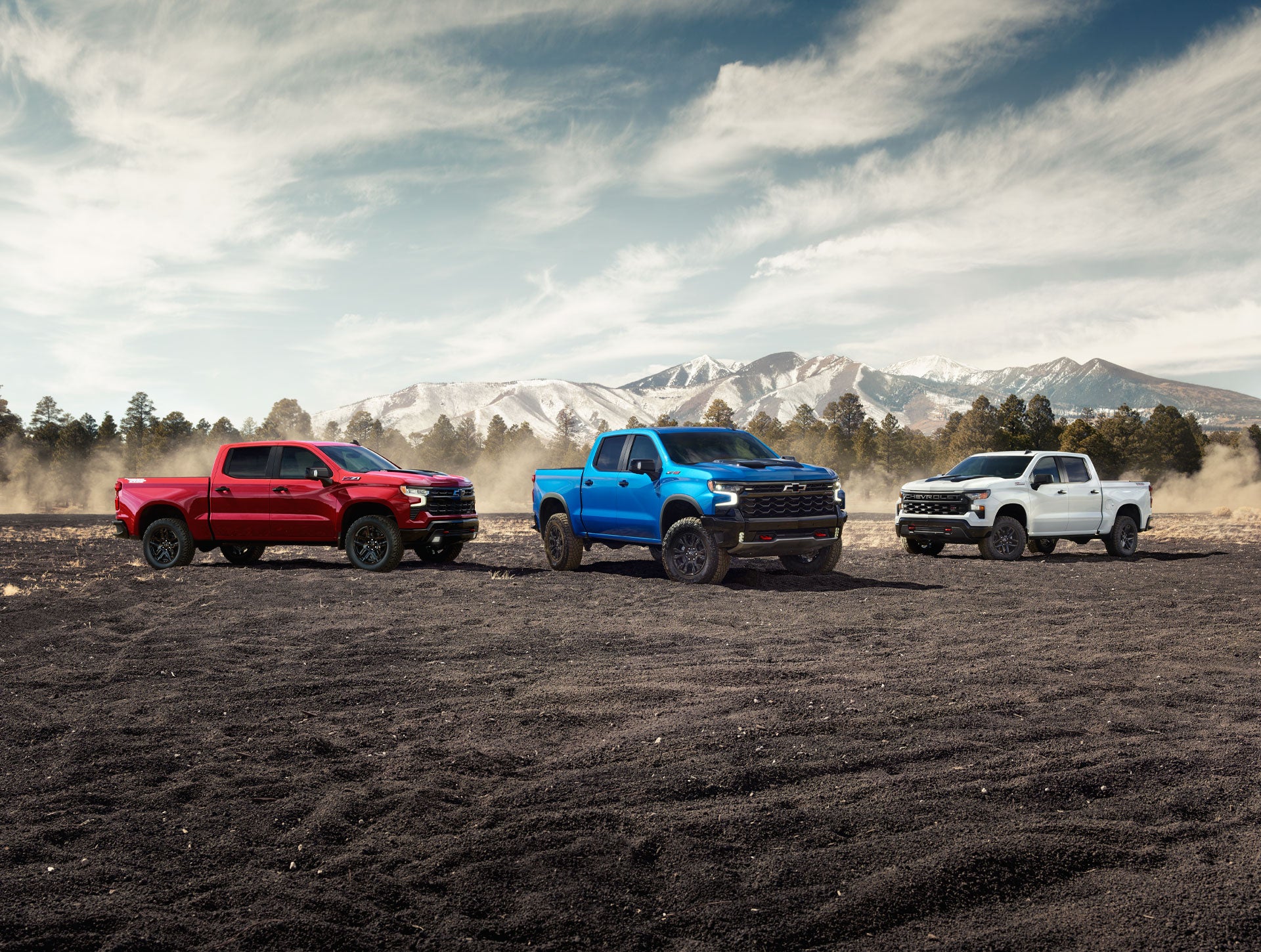 Three 2027 Chevrolet Silverado 1500 pickup trucks (red, blue, white) parked in a row on a dirt field with a mountain range backdrop.