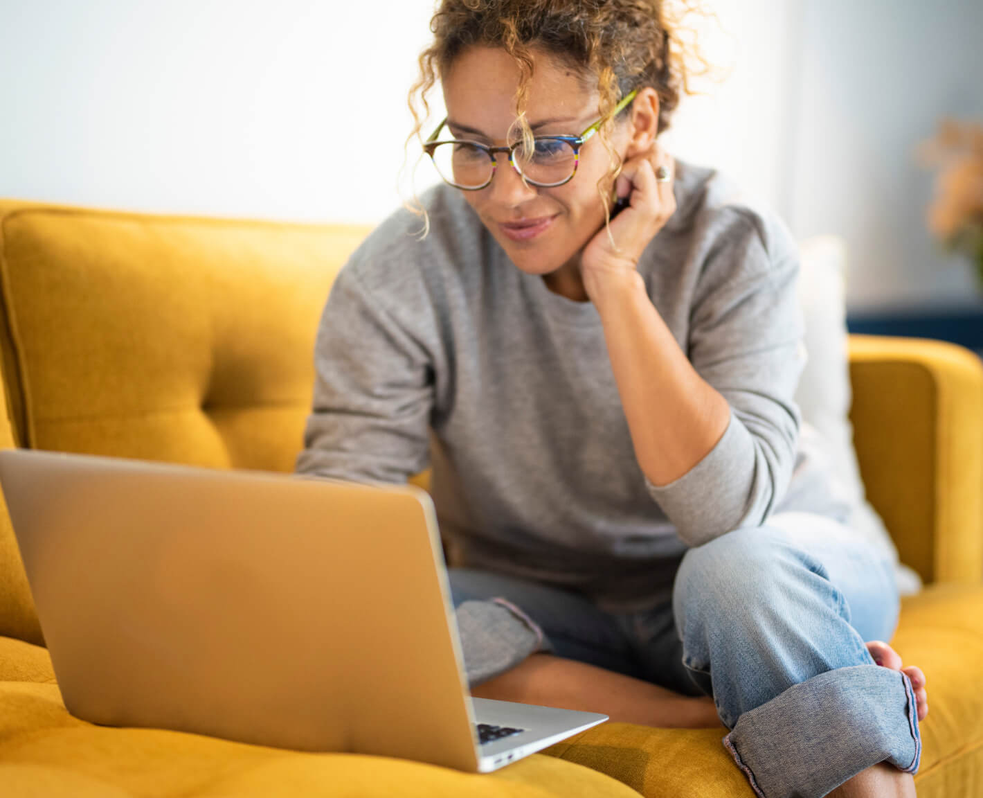 A woman sitting on a yellow couch smiles at her laptop.